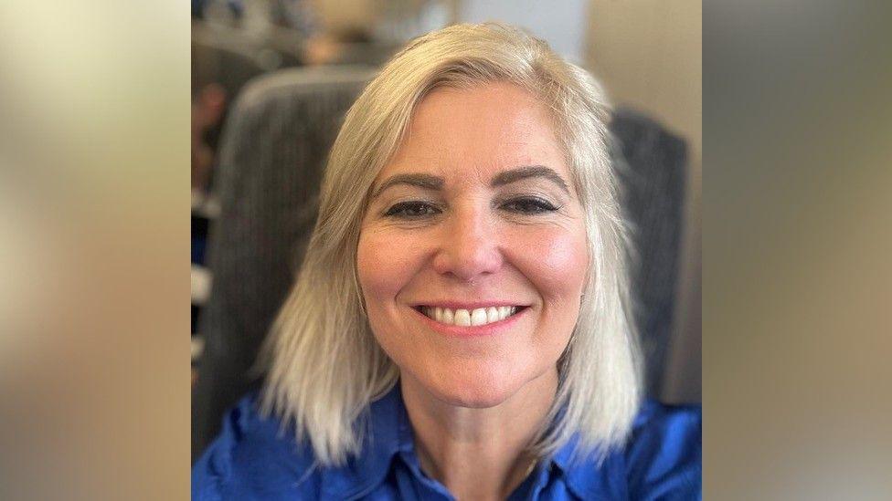 A selfie of Ms Catt, who appears to be on a train. She has a light blonde bob and is smiling at the camera. She is wearing a bright blue button-up shirt.