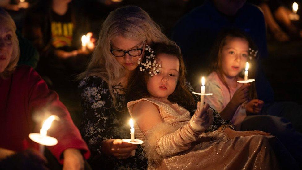 A woman and two daughters sit on the ground, holding candles