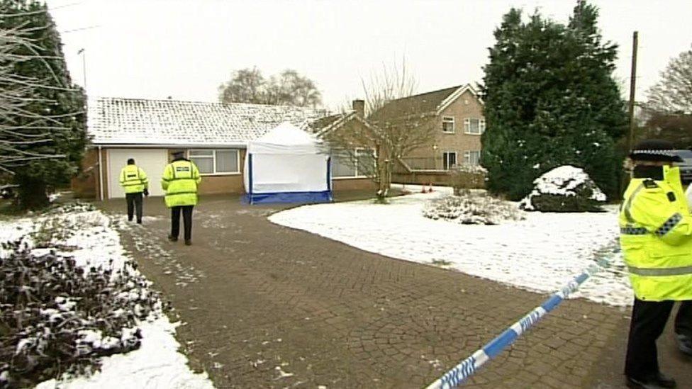 A snow-covered bungalow. A snow-covered garden and cobbled path are in front of the bungalow. There is police tape across the drive and three police officers, wearing yellow coats, are on the drive.