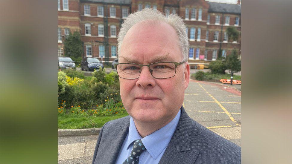 Simon Burwood who is wearing a blue suit and tie and wearing small rectangle glasses