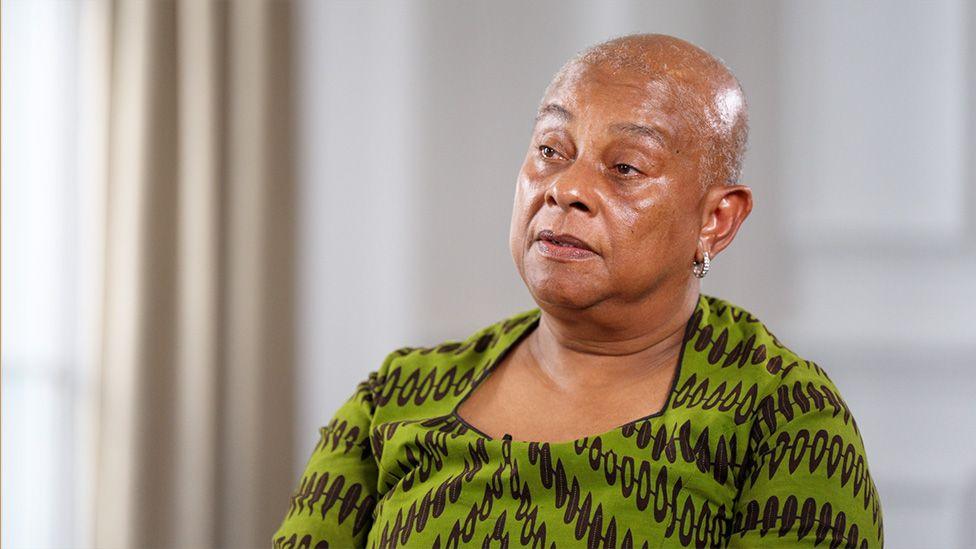 Doreen Lawrence wears a green dress with black patterning, and is sitting in a large formal room