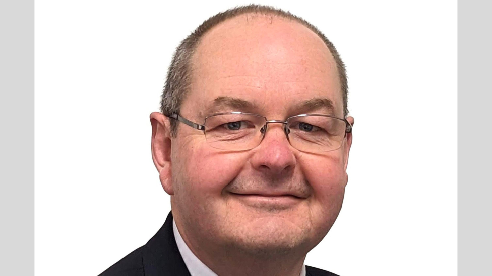 A smiling Chris Bally looking directly at the camera, in glasses and wearing a suit and shirt. 