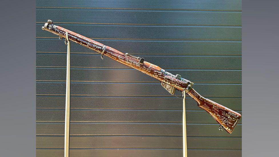 A decomissioned 1939 Lee-Enfield rifle on a stand against a metal door, its dark wood covered in white signatures