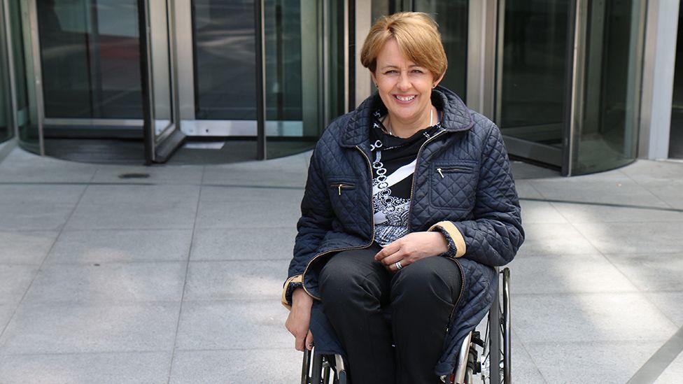 Baroness Tanni Grey-Thompson sitting in a wheelchair, wearing a navy blue quilted jacket and smiling. She has short, light brown hair. There are the revolving doors of an office entrance behind her.