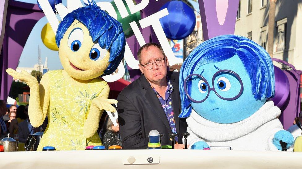 John Lasseter is pictured comically frowning in exaggerated sadness, seated between the characters of Joy and Sadness from Pixar's "Inside Out"