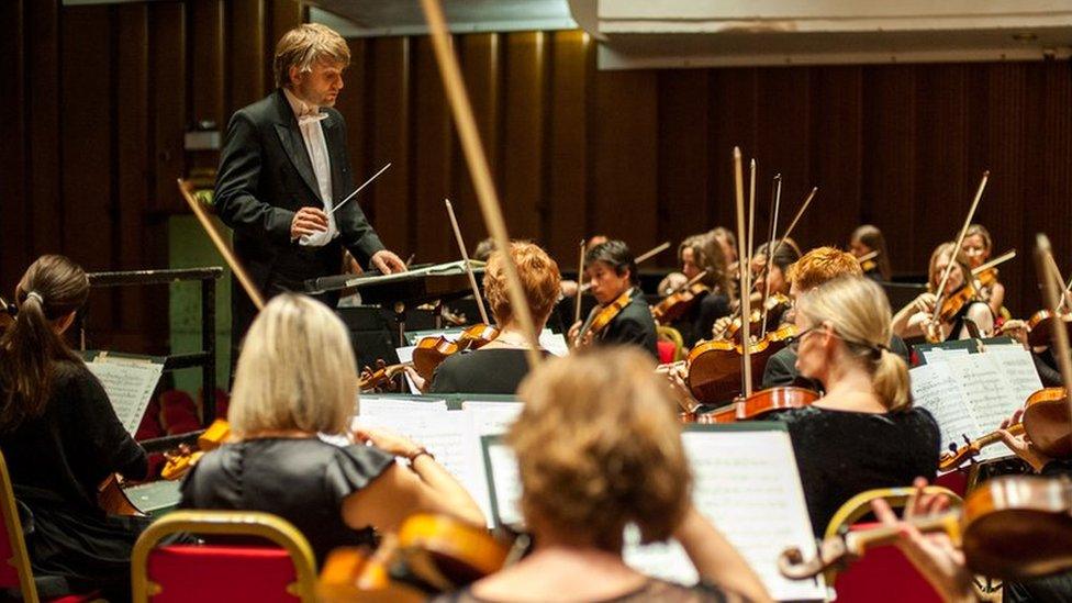 Kiril Karabits in formal dress and holding a conductors baton is standing facing an orchestra and conducting them while they play