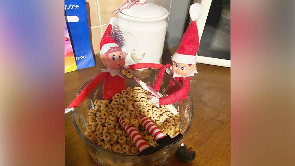 Two elves near a cereal bowl. One is sitting inside the bowl, the other leaning over it