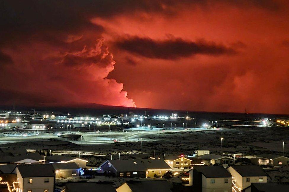 Houses in the village of Hafnarfjordur are seen with smoke billowing in the distance, and lava colours the night sky orange - following an volcanic eruption on the Reykjanes peninsula, western Iceland