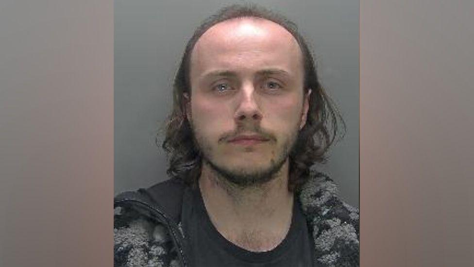 A custody photo of Samuel Robinson. He is facing the camera and has no expression on his face. He has a receding hairline with thin dark hair that comes to just above his shoulders. He is wearing a grey and black coat with a dark T-shirt on underneath.