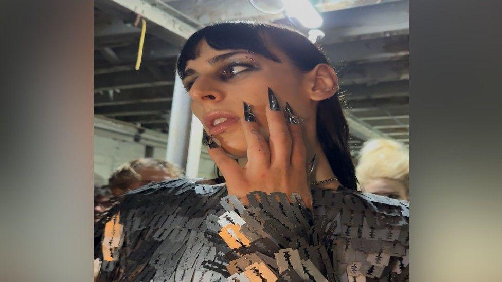 A headshot of a woman who is a model with a black fringe and black mid-length hair tucked back behind her ears. She is holding her hand on her face and she has long spiky, black nails. She is posing so is standing side on and looking into the distance.