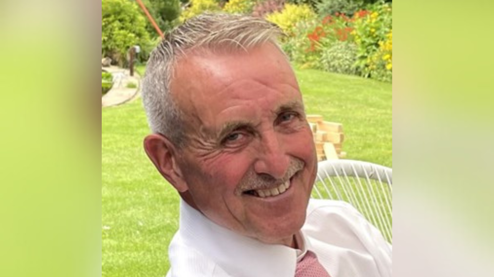 An older man with short grey hair, sitting in a garden, smiles at the camera.