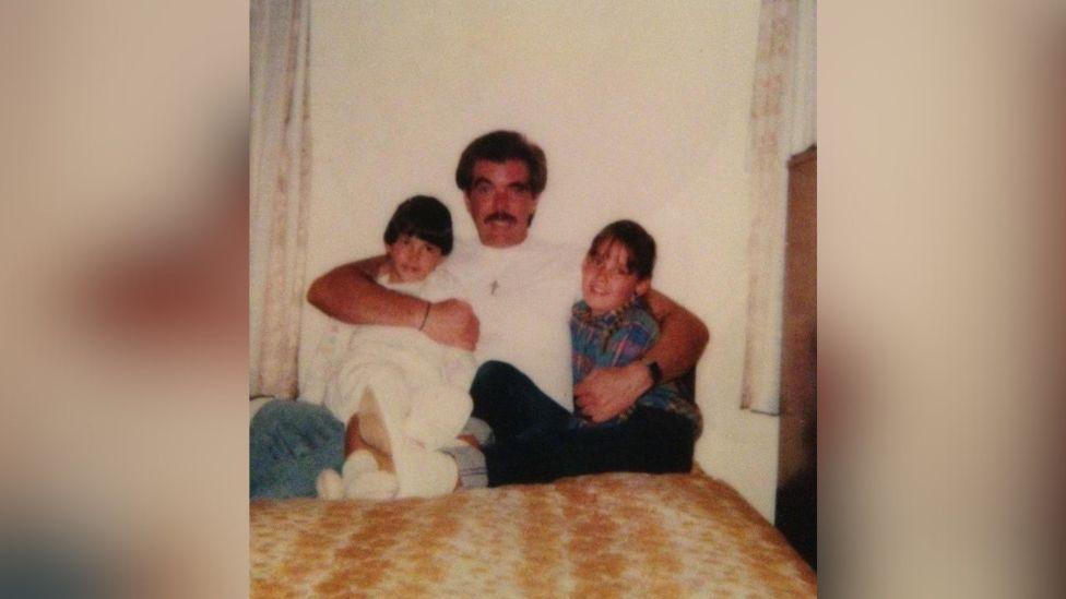 An old photograph of Tony and his two daughters. They are sat on a bed and he has his arms around them