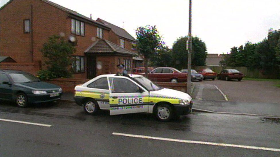 Police near Victoria Hall's home following her disappearance in 1999. A uniformed officer gets out of a police car parked on the side of the road. Houses and other parked cars can be seen behind on a side road.  