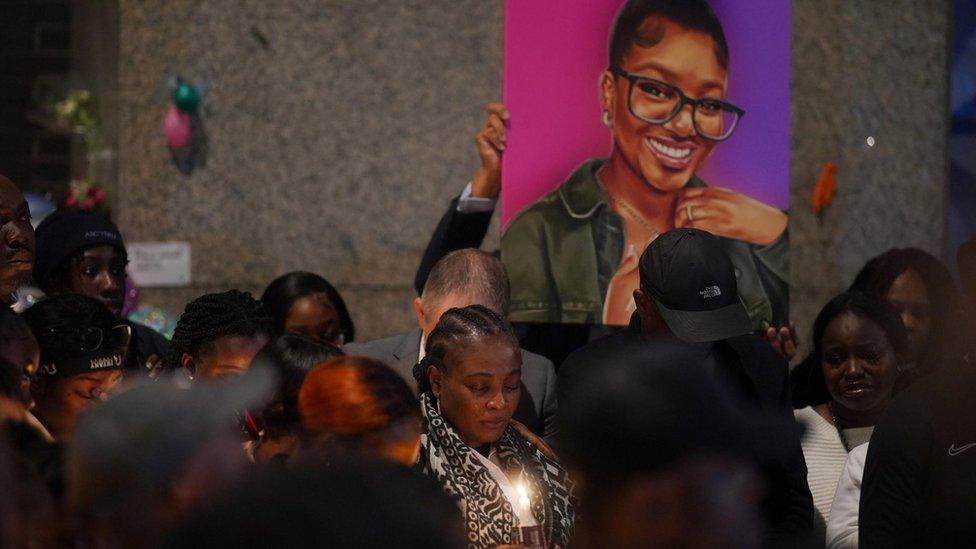 A woman holds a lit candle while surrounded by other people, one of whom is holding a painting of Elianne