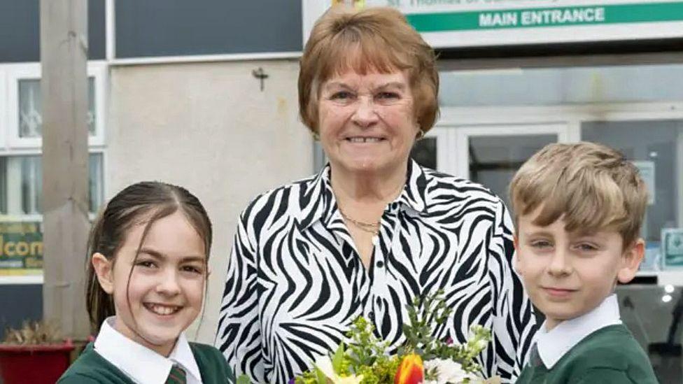 Mrs Hewlett stands outside the school with two children, they hold a large bouquet of flowers. She wears a zebra print shirt