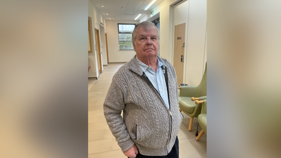 A man smiling at the camera in a hospital wearing a grey jacket and a baby blue shirt and black trousers