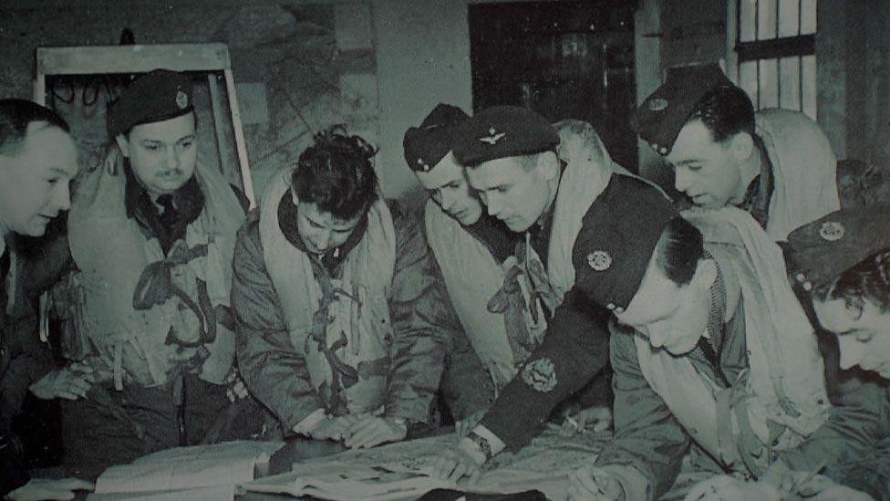 A black and white image of eight 1940s or 1950s RAF personnel. They are bending over a chart on a table and are wearing uniforms. Most of the men have life belts on over their uniforms.