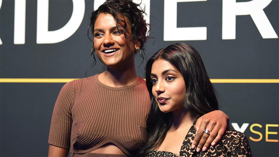 L-R Simone Ashley and Charithra Chandran. Simone is a 27-year-old British Asian woman with brown eyes and curly brown hair which is tied back. She looks to her right and smiles, wearing a ribbed brown top. Her left arm is around Charithra's shoulders. Charithra, a 27-year-old British Asian woman, stands about a foot shorter than Simone and wears a square-necked leopard print top. She has brown eyes and long brown hair which is worn loose down her back. The actresses are pictured at an event in front of a dark grey backdrop, dissected by a yellow horizontal line with large white print above it.