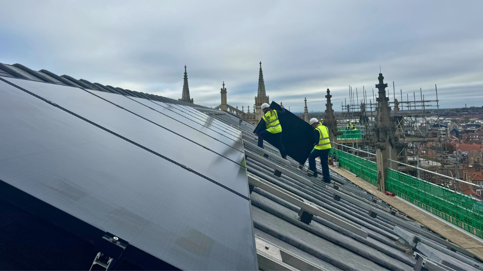 Two people in hi-vis jackets and white helmets carrying a solar panel onto the slate roof of York Minster 