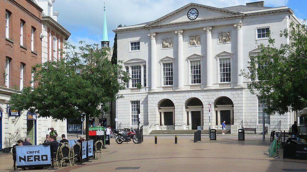 Shire Hall in Chelmsford, at the top of High Street. It is a grand, white building with pillars, doors, windows and a clock on its facade. To the left are people sat outside a Caffe Nero and to the right is a tree.