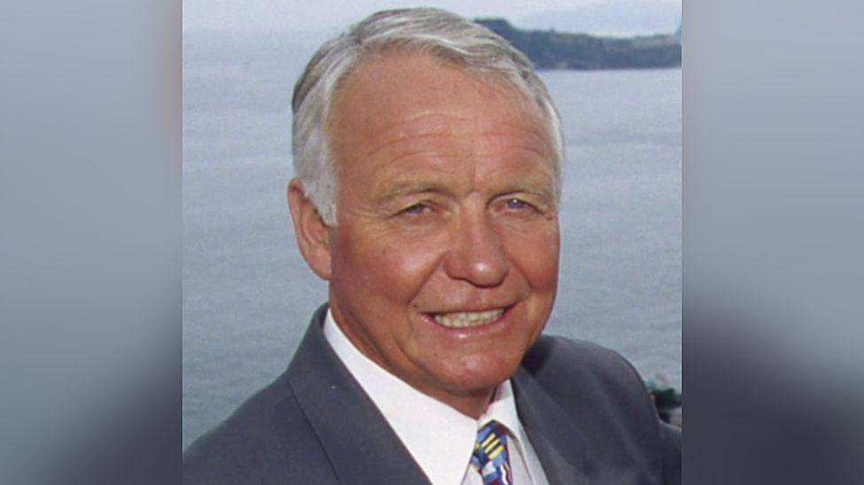 Craig Rich seen wearing a grey suit, white shirt and patterned tie. He is pictured with the sea in the background.