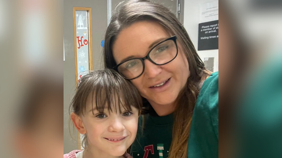 Ashley Wiseman wearing a green jumper and black glasses while standing in front of a door in hospital. She is with Isla, who is wearing a pink jumper. Both are smiling.