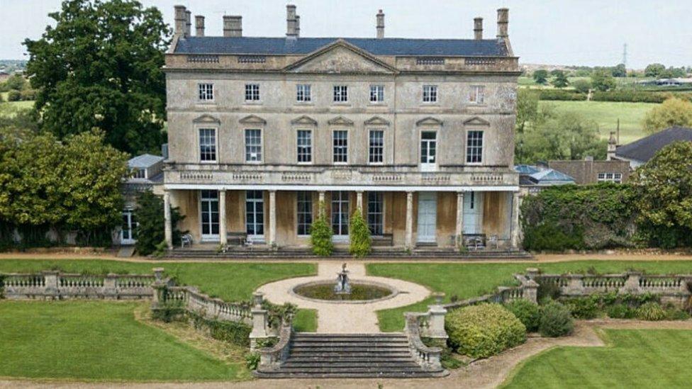 A large period building with stone pillars, surrounded by gardens and hedgerows, seen from the air