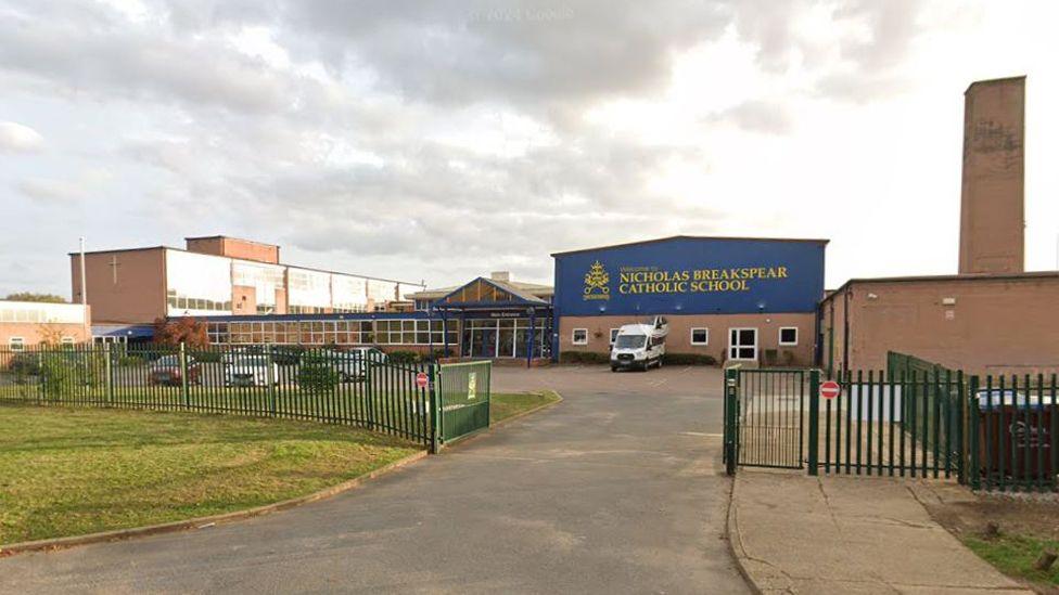 A school building seen from outside the school gates. It has green metal fences. The main building is partly blue with yellow writing, reading "Nicholas Breakspear". The rest of the building is largely made up of windows.