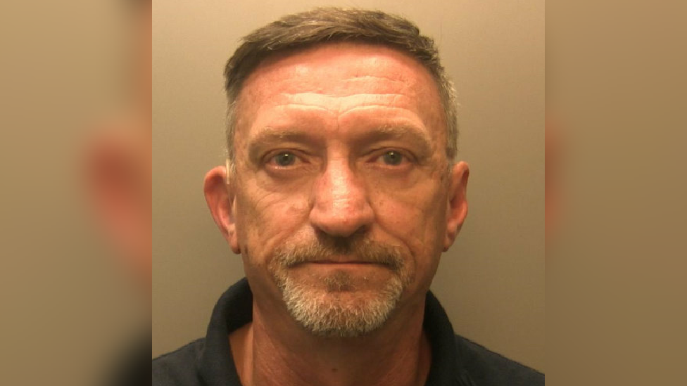 A mugshot of an elder man, looking towards the camera lens. He has grey hair, moustache and a beard. He is wearing a navy blue polo shirt. Stood against a white backdrop.