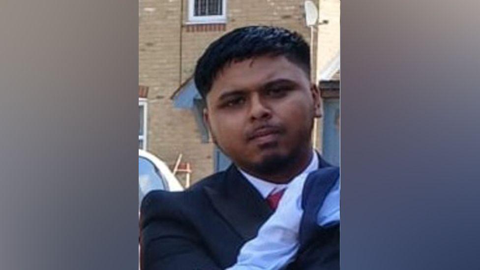 Yusef looking straight at the camera, dressed smartly as though going to work or a wedding. He is wearing a dark suit jacket, a white shirt and red tie. He is standing in front of a brick house and has a light goatee beard and moustache, and slicked short dark hair.