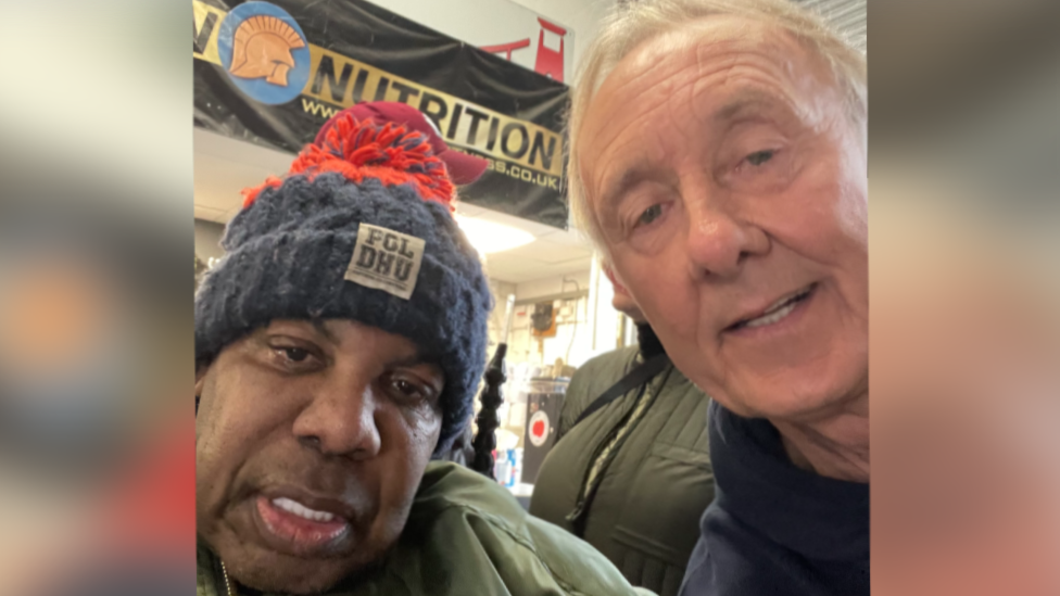 David 'Syd' Lawrence and Andy Brassington. David 'Syd' Lawrence is on the left of the frame wearing a blue and red bobble hat. Andy Brassington is next to him. He has short white hair and is leaning in towards Mr Lawrence, looking at the camera and smiling. 