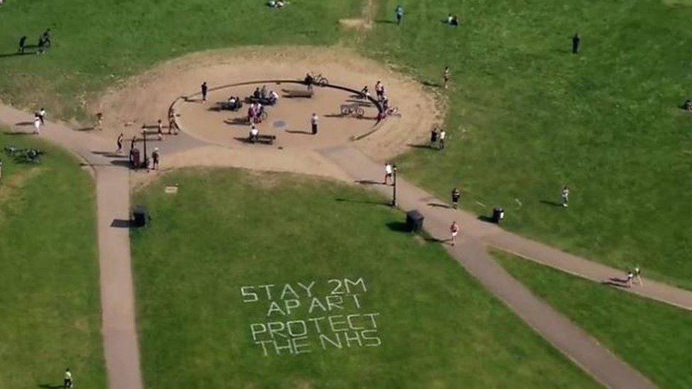 Lockdown in a park, with sign written on grass saying 'stay two metres apart, protect the NHS'