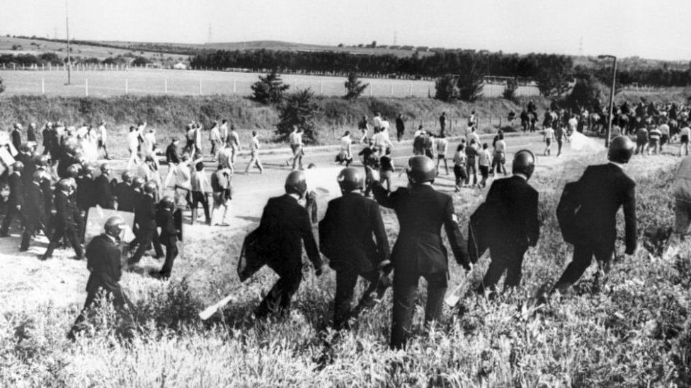 A black and white image of miners and police officers with riot gear walking along a field. 