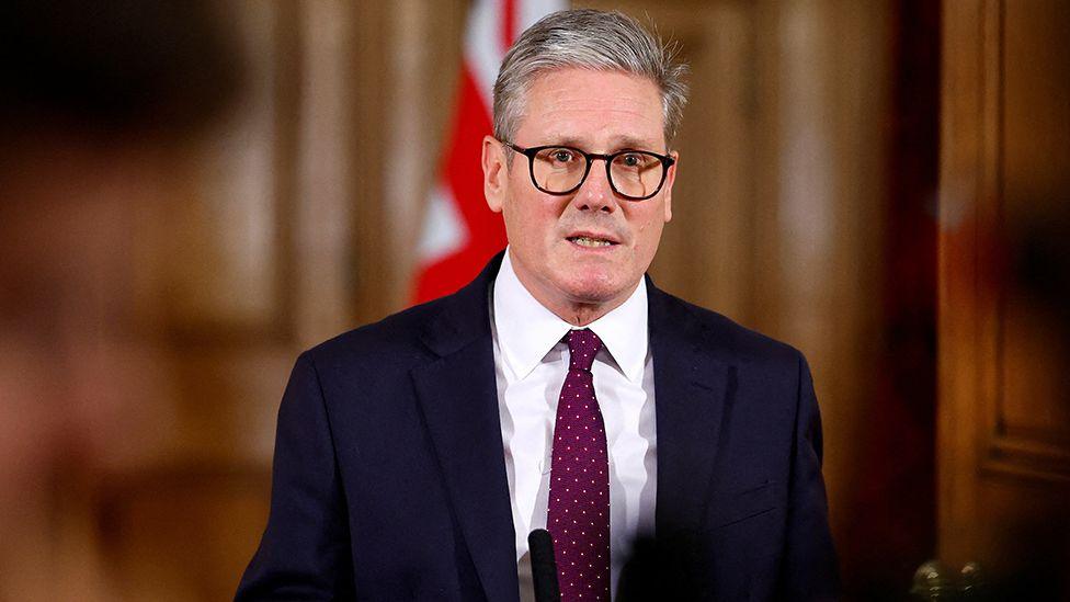 Prime Minister Keir Starmer looks at the camera in a suit, purple tie, and glasses, as he delivers a statement on conflict in the Middle East inside Downing Street on 1 October