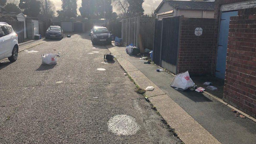 A residential road where three cars are parked. Litter blown from recycling sacks has been blown across the street due to the sacks being spilt.