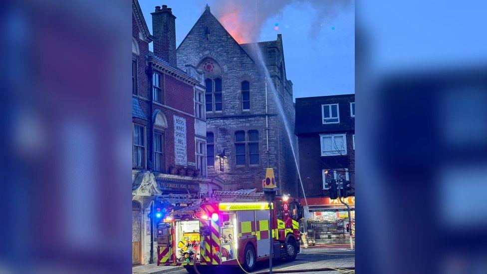 A three-story stone building with a pointed roof, with the orange glow of fire at the top. A fire engine is parked in front of it, with a jet of water aimed at the flames.