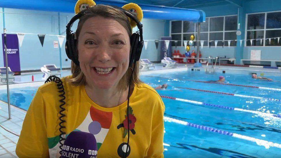 Woman with blond hair wearing headphones with yellow bear ears with a yellow T-shirt, poppy and purple microphone and swimming pool behinf