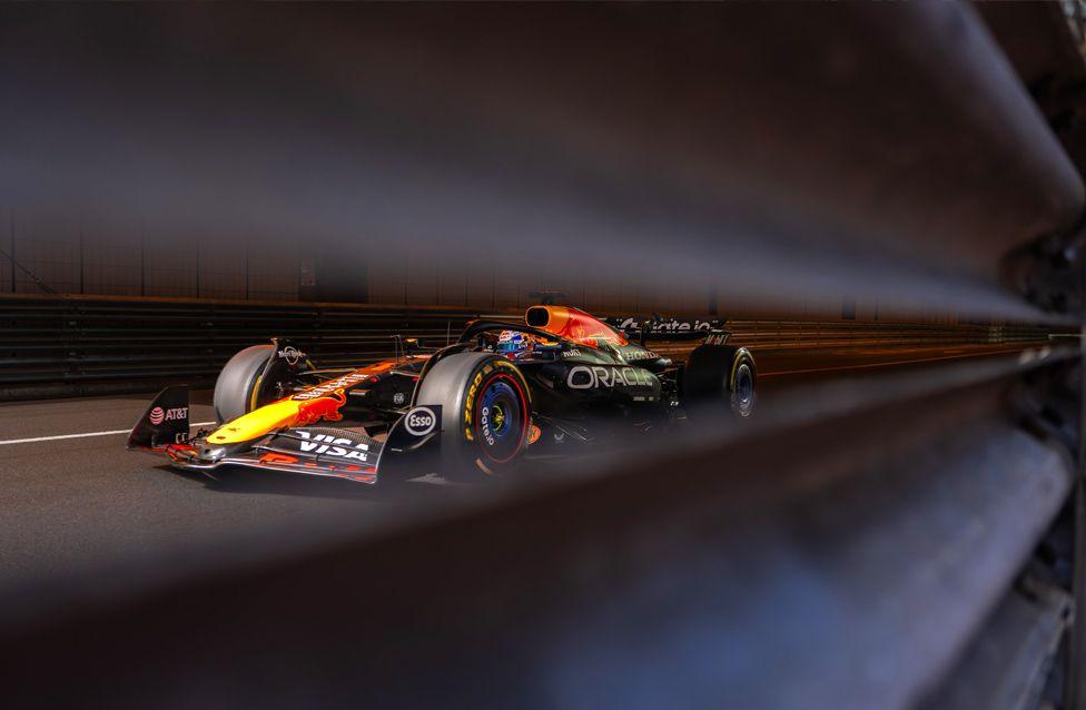 A Red Bull Formula 1 car speeds through a tunnel, captured through the slats of a metal barrier. The car features vibrant red, yellow, and black colours with visible sponsor logos.