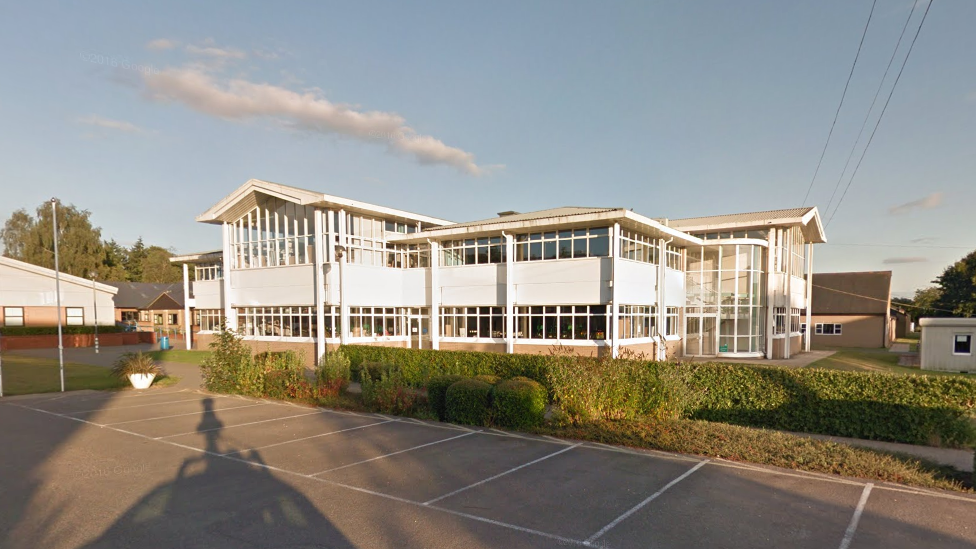 Wymondham College, in Norwich: a white two-storey building, with large glass-panelled windows running continuously on each floor. In front of the building is a green hedge which separates it from the car park. 