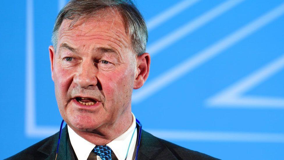 A close-up of Rupert Lowe. He has short grey hair and is looking to his right while giving a speech. He is wearing a grey suit jacket over a white shirt and blue tie. Behind him there is a blue coloured screen.