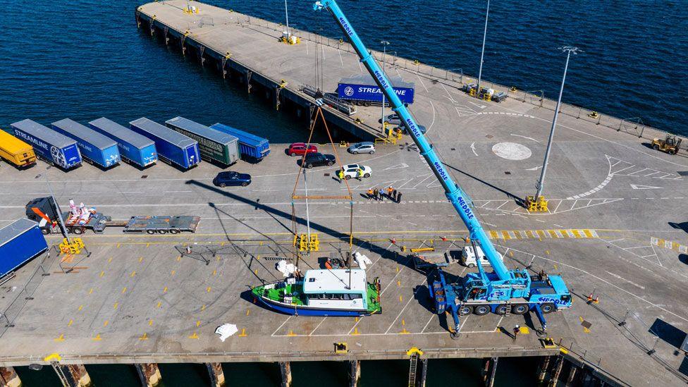 A small ferry viewed from above being lifted by a crane on dock at harbour