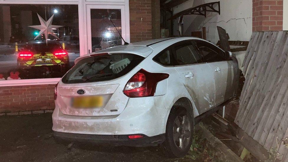 A white Ford Focus is wedged in the side of a white bungalow. Damage to the outside of the bungalow can be seen, and a wooden fence is now at an angle with some of its vertical panels broken. In the reflection of the neighbouring hair salon window a police car and its red rear lights can also be seen. 