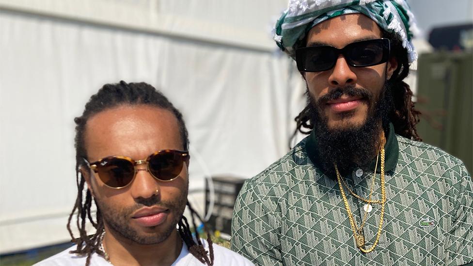 Drummer Bobbie (with an ie) on the left and frontman Bobby (with a y) on the right. Both are wearing sunglasses and Bobbie with an ie has a white T-shirt on while Bobby with a y has a green and white shirt on and is wearing gold chains around his neck