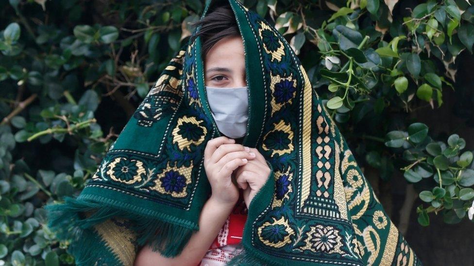 A young Palestinian wearing a face mask holds a prayer mat over their head