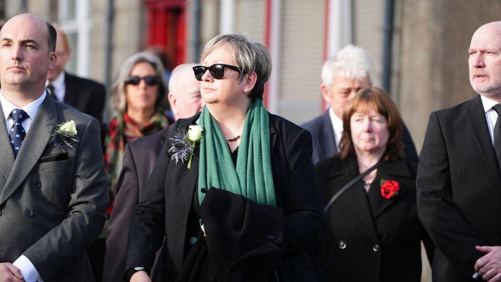 Joanna Cherry, wearing a dark suit, green scarf and dark sunglasses stands among mourners outside a church 