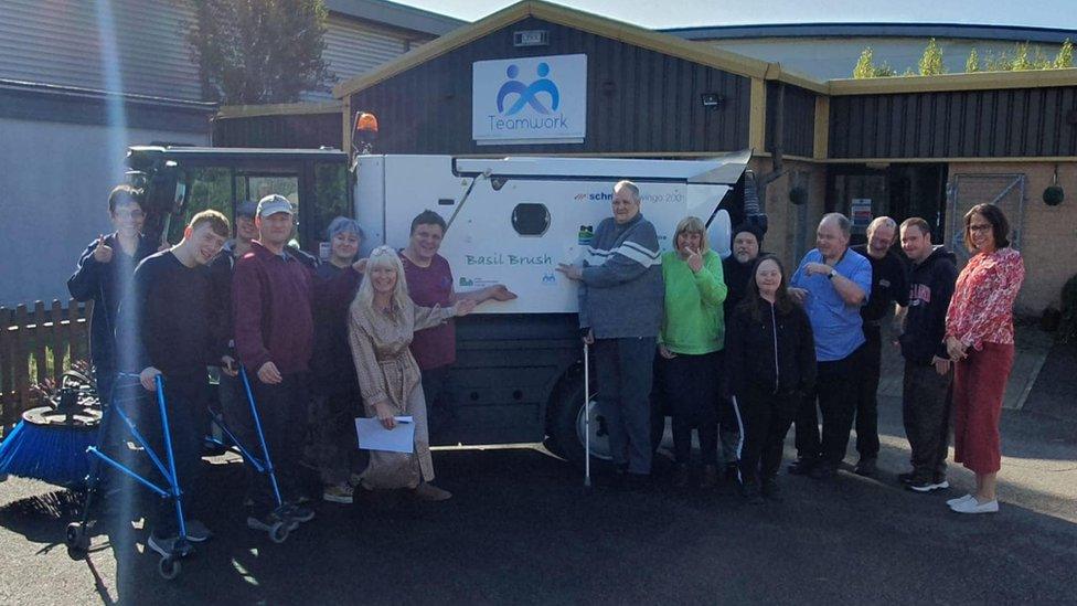 A group of 14 people of varying ages stand in front of a street sweeper. One is pointing to the "Basil Brush" name.