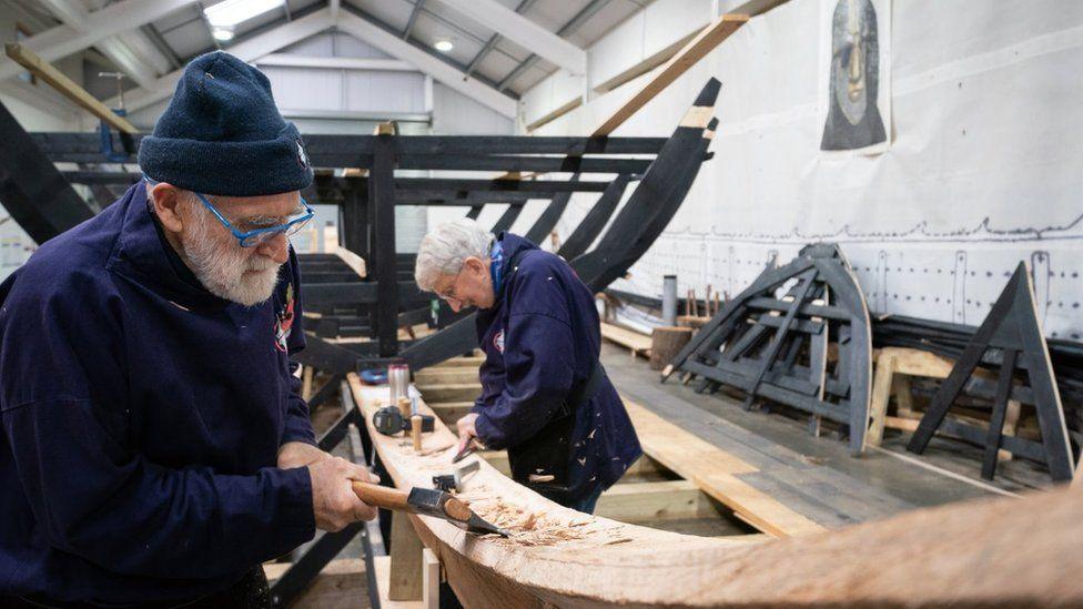 A gentleman is pictured close up chopping a wooden panel with a small axe. He has a short grey beard and is wearing a navy beanie hat with a navy jumper. Behind him on the opposite side of the panel another volunteer works the wood.