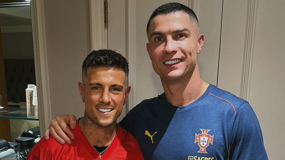 Cal Newsome has brown hair and is wearing a red top with black and white writing that reads "form". He has a silver cross necklace on. Cristiano has his arm around him, and has short black hair. He is wearing a navy t-shirt with the puma log on it, and the Portugal logo