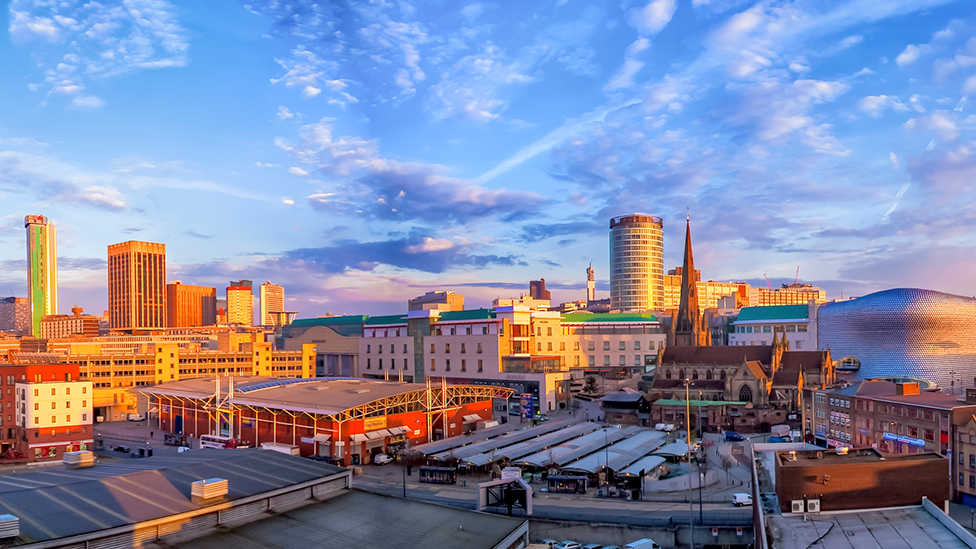 The cityscape of Birmingham showing industrial buildings in the foreground and high-rise towers on the horizon.