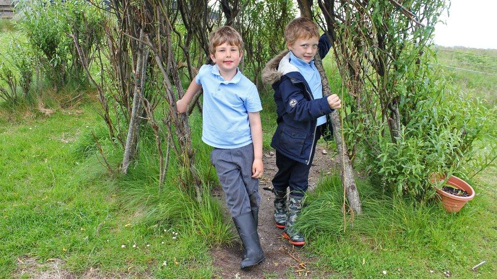 Two boys stand in a den made out of willow branches.